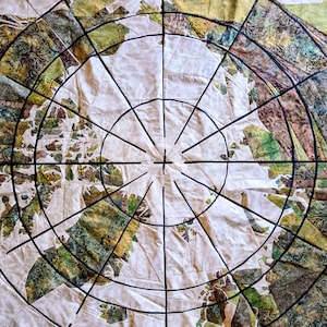 May include: A close-up of a quilt with a circular design. The quilt features a cream-colored center with radiating black lines and sections of green, brown, and yellow patterned fabric. The overall design resembles a compass or a map.