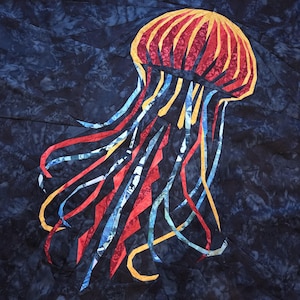 May include: A fabric jellyfish design on a dark blue background. The jellyfish has a red and yellow head with long, flowing tentacles in red, blue, and yellow. The artwork is likely a quilt or textile art piece.