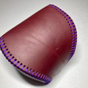 May include: A burgundy leather cuff with purple stitching. The cuff is curved and appears to be handmade. The stitching is evenly spaced and adds a decorative element to the leather. The cuff is likely designed to be worn on the wrist or forearm.
