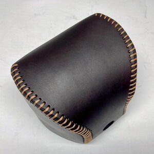 May include: A dark brown leather gear shift knob cover featuring a smooth, matte finish and prominent, light tan baseball-style stitching along the curved edges. The cover is shaped to fit over a standard automotive gear shifter handle.