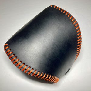May include: A curved black leather sleeve with orange stitching. The orange thread provides a striking contrast. The item appears to be hand-stitched. The sleeve is likely for a small item.