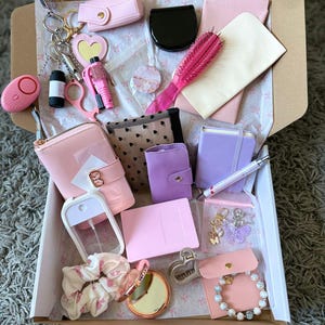 May include: A selection of pink and purple accessories, such as a wallet, keychain, brush, compact mirror, and scrunchie, are arranged inside a cardboard box. Other items include a small notebook, pen, and a bracelet. The items are on a white paper with pink bows.