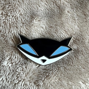 Könnte beinhalten: Eine schwarze und weiße Emaille-Anstecknadel mit einem Cartoon-Waschbären mit blauen Augen. Die Anstecknadel hat eine silberne Oberfläche.