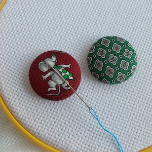 May include: A close-up of two fabric buttons being sewn onto a white fabric. One button features a red background with a white mouse illustration. The other button has a green background with a repeating pattern of grey and yellow squares.