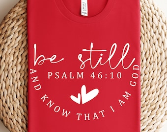 May include: A red t-shirt with the words "be still" in a cursive font, along with "Psalm 46:10" and "and know that I am God" in a circular arrangement. Two white heart shapes are below the text. The shirt is displayed on a woven, circular mat.