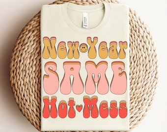 Op de afbeelding: Een crèmekleurig t-shirt met de tekst "New Year Same Hot Mess" in retro-letters. De tekst is in tinten oranje, roze en rood. Het shirt is opgevouwen op een geweven, ronde placemat.