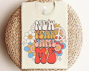 Op de afbeelding: Een licht beige T-shirt met een retro grafisch ontwerp met de tekst "New Year Same Me" in kleurrijke, gestileerde letters omringd door bloemen en een vredesteken.