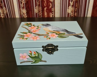 Puede incluir: Una caja rectangular de madera de color azul claro con una ilustración pintada a mano de un pájaro posado en una rama florida en la tapa y una rama floral más pequeña en el panel frontal. Incluye un cierre decorativo de metal bronce.