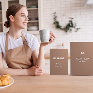 May include: A woman in a white t-shirt and brown apron holds a mug in a kitchen setting. Two brown rectangular objects are on the table, one labeled "US TRADE 6 x 9 inches" and the other "A4 8.27x11.69 inches". A plate of croissants is on the table.