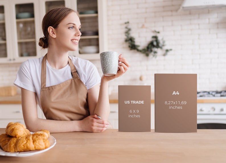 May include: A woman in a white t-shirt and brown apron holds a mug in a kitchen setting. Two brown rectangular objects are on the table, one labeled "US TRADE 6 x 9 inches" and the other "A4 8.27x11.69 inches". A plate of croissants is on the table.