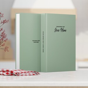 May include: Two sage green books with the text "YOUR FRONT TEXT Goes Here", "YOUR SPINE TEXT GOES HERE", and "YOUR BACK TEXT GOES HERE". A white vase with red flowers and a red and white checkered napkin are in the foreground. The books are on a white surface.