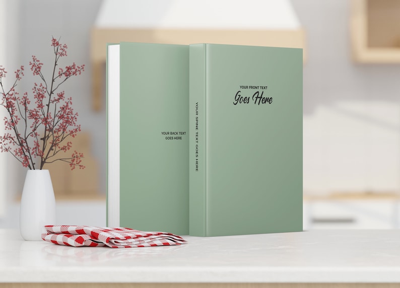 May include: Two sage green books with the text "YOUR FRONT TEXT Goes Here", "YOUR SPINE TEXT GOES HERE", and "YOUR BACK TEXT GOES HERE". A white vase with red flowers and a red and white checkered napkin are in the foreground. The books are on a white surface.