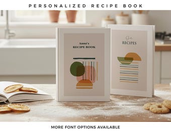 May include: Two hardcover recipe books with white spines and covers featuring beige panels. The left book displays the text Anna's RECIPE BOOK above abstract shapes in green, orange, and blue. The right book shows the text RECIPES above similar abstract shapes.