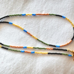May include: A beaded necklace featuring a repeating pattern of small, round seed beads in black, olive green, bright orange, yellow, and light blue. The necklace is finished with a gold-toned metal lobster claw clasp and a small circular jump ring.