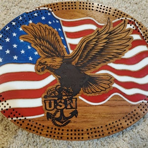 May include: A wooden oval-shaped plaque with a red, white, and blue American flag design. A bald eagle is flying over the flag with its wings spread. The eagle is holding an anchor in its talons. The words "USN" are printed below the anchor.