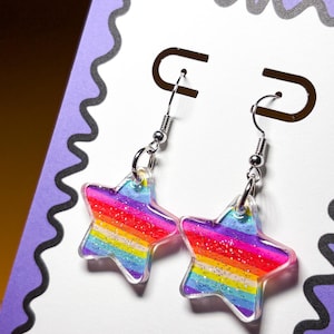 May include: A pair of star-shaped earrings with a rainbow striped design. The earrings have a clear, glittery finish and silver-colored hooks. The earrings are displayed on a white card with a purple border.