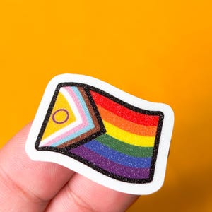 May include: A colorful sticker featuring a rainbow flag design with additional symbols. The sticker has a white border and is held against a bright orange background. The rainbow flag includes red, orange, yellow, green, blue, and purple stripes.