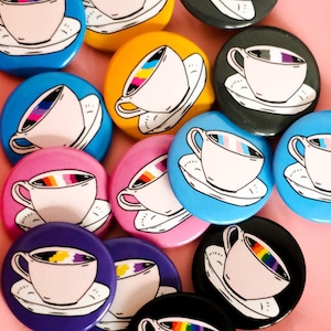 May include: A collection of colorful round buttons with a teacup design. Each button features a white teacup and saucer with a rainbow-colored liquid inside. The buttons are in various colors, including blue, yellow, pink, and black, against a pink background.