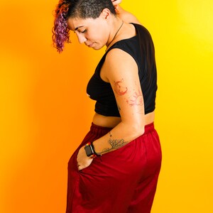 May include: A person wearing a black sleeveless top and burgundy shorts. The person has a tattoo on their arm and is wearing a smartwatch. The background is a gradient of yellow and orange.