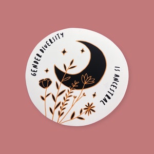 May include: A round sticker with a black crescent moon, stars, and floral elements in gold and black. The text "GENDER DIVERSITY IS ANCESTRAL" is printed around the edge of the sticker on a white background. The sticker is on a pink background.