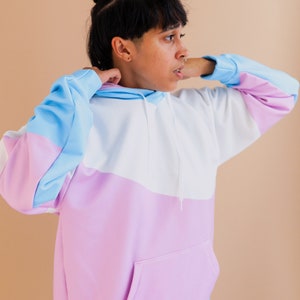 May include: A color-block hoodie in pastel shades of blue, white, and pink. The hoodie features a front pocket, a drawstring hood, and long sleeves. The sleeves and hood are trimmed in blue, and the body of the hoodie is white and pink.