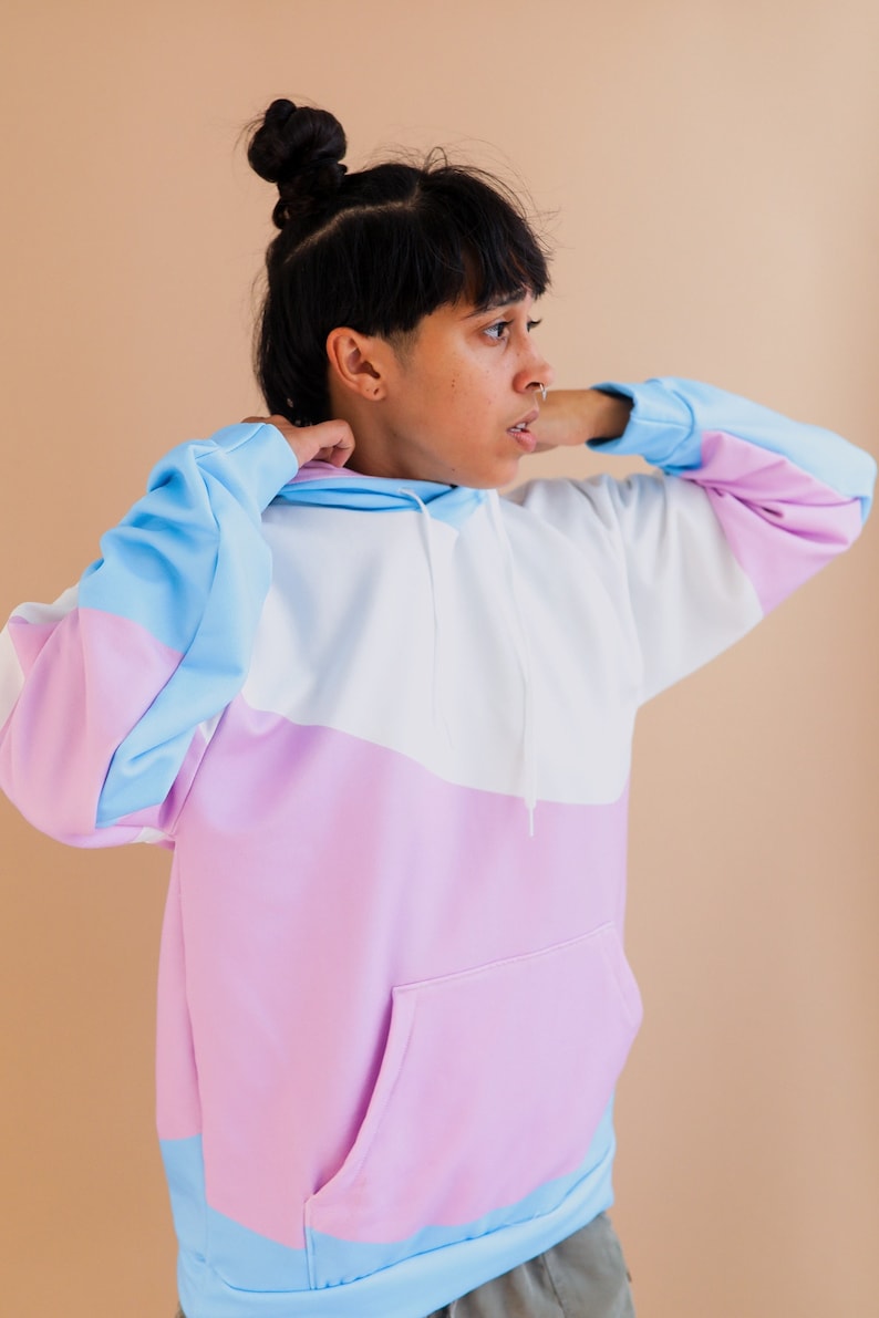 May include: A color-block hoodie in pastel shades of blue, white, and pink. The hoodie features a front pocket, a drawstring hood, and long sleeves. The sleeves and hood are trimmed in blue, and the body of the hoodie is white and pink.