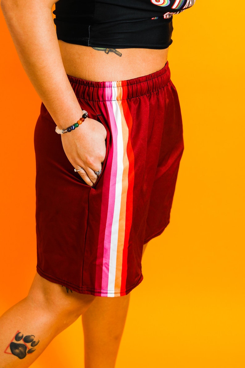 May include: Burgundy athletic shorts with a colorful vertical stripe down the side. The stripe features bands of white, pink, orange, and red. The shorts have an elastic waistband and pockets.