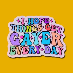 May include: A colorful sticker with the text "I HOPE THINGS GET GAYER EVERY DAY" in a retro font. The sticker has a glittery finish and is set against a mustard yellow background. The text is in various colors, including pink, blue, and orange.