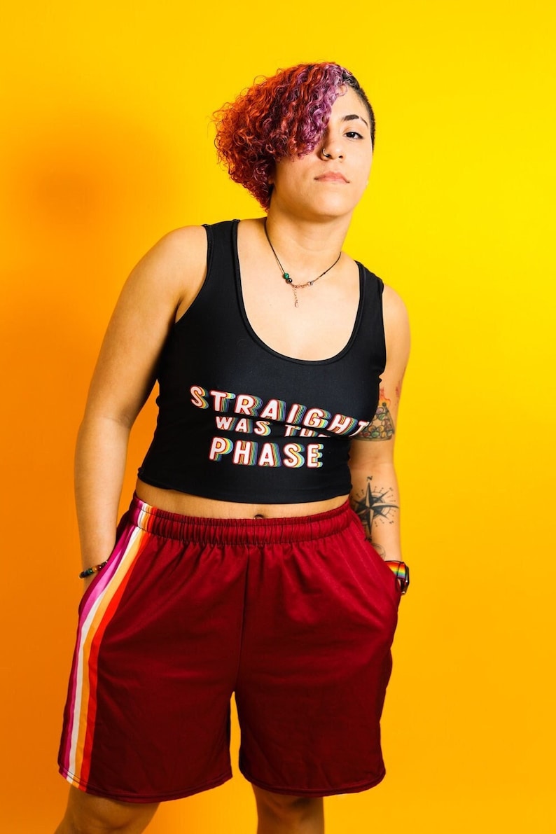 May include: Black crop top with the text "STRAIGHT WAS THE PHASE" in rainbow colors. The model is wearing burgundy shorts with a rainbow stripe down the side. The background is a solid yellow.