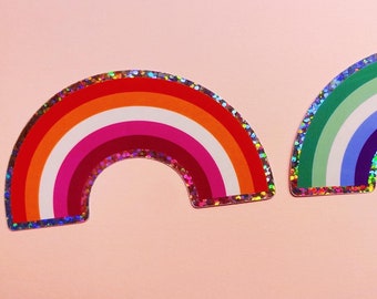 May include: Two rainbow-shaped stickers with glitter edges. One features the colors of the lesbian pride flag: orange, red, pink, white, and dark pink. The other has green, white, blue, and purple stripes.