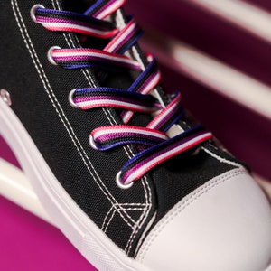 Pode incluir: T&ecirc;nis de lona preto de cano alto com costura branca e biqueira branca. Os cadar&ccedil;os s&atilde;o listrados em vermelho, branco, roxo e azul. O t&ecirc;nis est&aacute; sobre um fundo rosa e branco.
