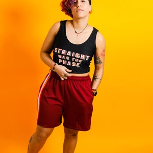 May include: A person wearing a black tank top with the text "STRAIGHT WAS THE PHASE" in rainbow colors, paired with maroon shorts with a white and orange stripe. The background is a vibrant orange.