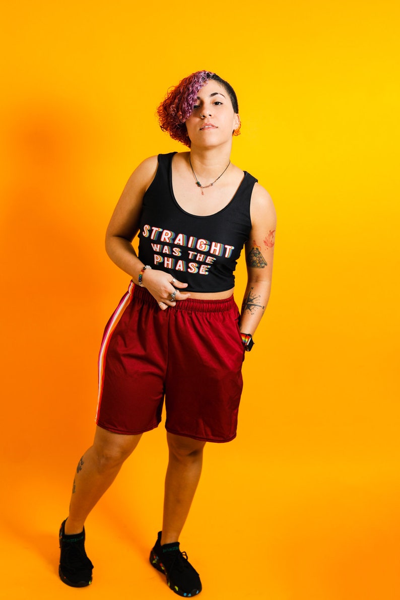 May include: A person wearing a black tank top with the text "STRAIGHT WAS THE PHASE" in rainbow colors, paired with maroon shorts with a white and orange stripe. The background is a vibrant orange.