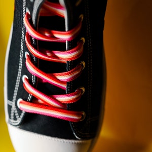 Pode incluir: Close-up de um t&ecirc;nis de lona preto com biqueira branca e cadar&ccedil;os listrados coloridos em tons de vermelho, rosa e branco. O t&ecirc;nis est&aacute; sobre um fundo amarelo brilhante.