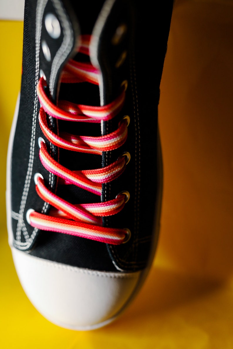 Pode incluir: Close-up de um t&ecirc;nis de lona preto com biqueira branca e cadar&ccedil;os listrados coloridos em tons de vermelho, rosa e branco. O t&ecirc;nis est&aacute; sobre um fundo amarelo brilhante.
