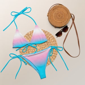 Puede incluir: Un conjunto de bikini de dos piezas con un diseño degradado en tonos rosa, blanco y azul. La parte superior es de estilo triangular con tirantes anudados, y la parte inferior es de corte clásico con lazos laterales. Una bolsa de paja tejida, gafas de sol y una esterilla tejida también están en la imagen.
