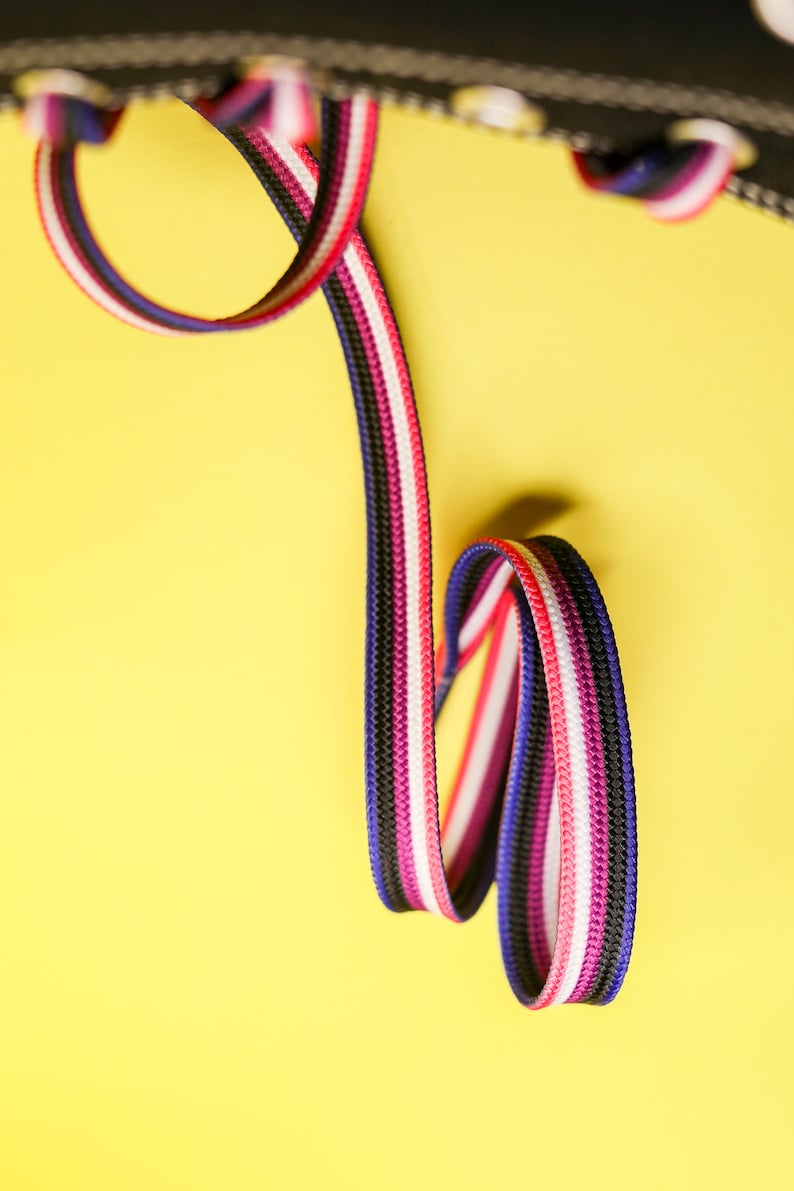 Pode incluir: Close-up de um sapato preto com atacadores coloridos. Os atacadores s&atilde;o uma mistura de preto, branco, rosa, vermelho e azul. O fundo &eacute; amarelo brilhante.