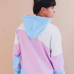 May include: A color-block hoodie in pastel shades of blue, white, and lavender. The hoodie features a drawstring hood and long sleeves. The design is a modern, casual style, suitable for everyday wear.
