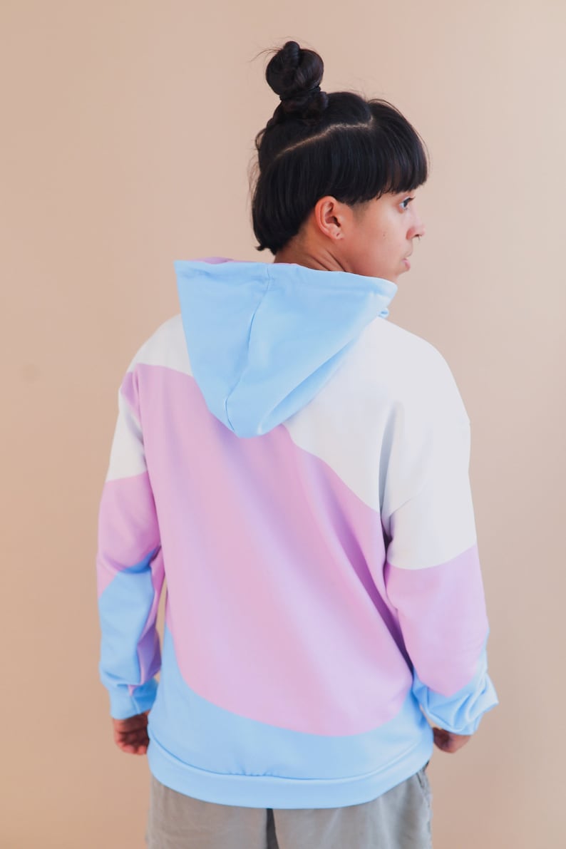 May include: A color-block hoodie in pastel shades of blue, white, and lavender. The hoodie features a drawstring hood and long sleeves. The design is a modern, casual style, suitable for everyday wear.