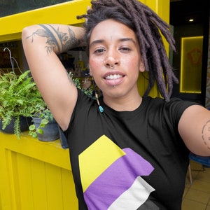 以下が含まれることがあります： 黒いTシャツ。黄色、紫、白、黒のノンバイナリープライドフラッグのデザイン。画像内の人物はTシャツを着ており、ドレッドヘアとタトゥーがあります。背景は黄色の壁です。