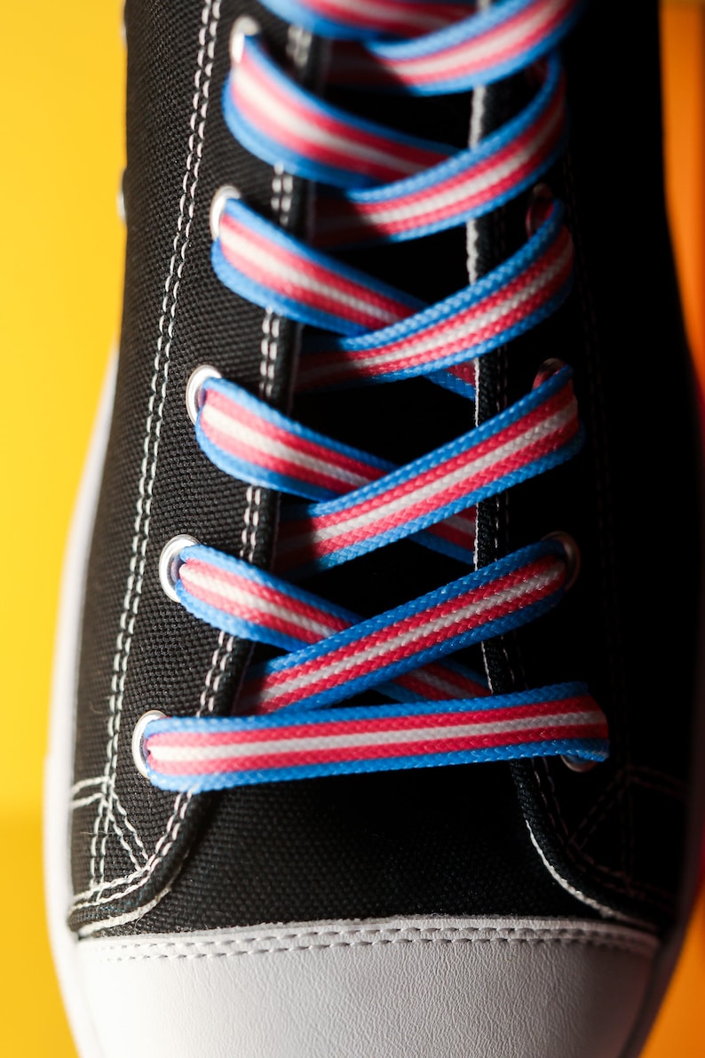 Pode incluir: Close-up de um t&ecirc;nis de lona preto com costuras brancas e biqueira branca. Os cadar&ccedil;os s&atilde;o listrados em azul, branco e rosa, criando um contraste colorido com o t&ecirc;nis preto. Os cadar&ccedil;os s&atilde;o enfiados em ilh&oacute;s prateados.