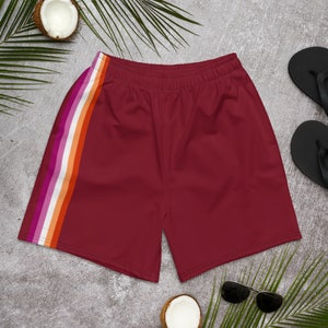 May include: Maroon athletic shorts with a side stripe in the colors of the lesbian pride flag: pink, orange, white, and orange. The shorts have an elastic waistband. The image also includes sunglasses, flip-flops, and coconuts.