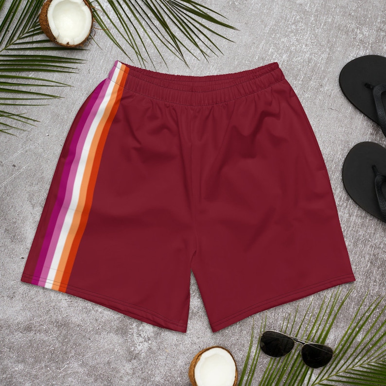 May include: Maroon athletic shorts with a side stripe in the colors of the lesbian pride flag: pink, orange, white, and orange. The shorts have an elastic waistband. The image also includes sunglasses, flip-flops, and coconuts.