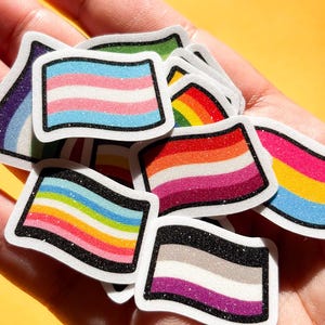 May include: A collection of small, flag-shaped stickers with various LGBTQ+ pride flag designs. The stickers feature rainbow, transgender, lesbian, asexual, and pansexual flag patterns, outlined in black with a glittery border. The background is a solid yellow.