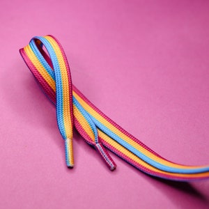 Pode incluir: Um par de atacadores listrados em rosa, amarelo e azul. Os atacadores s&atilde;o exibidos sobre um fundo rosa. Um acess&oacute;rio vibrante.