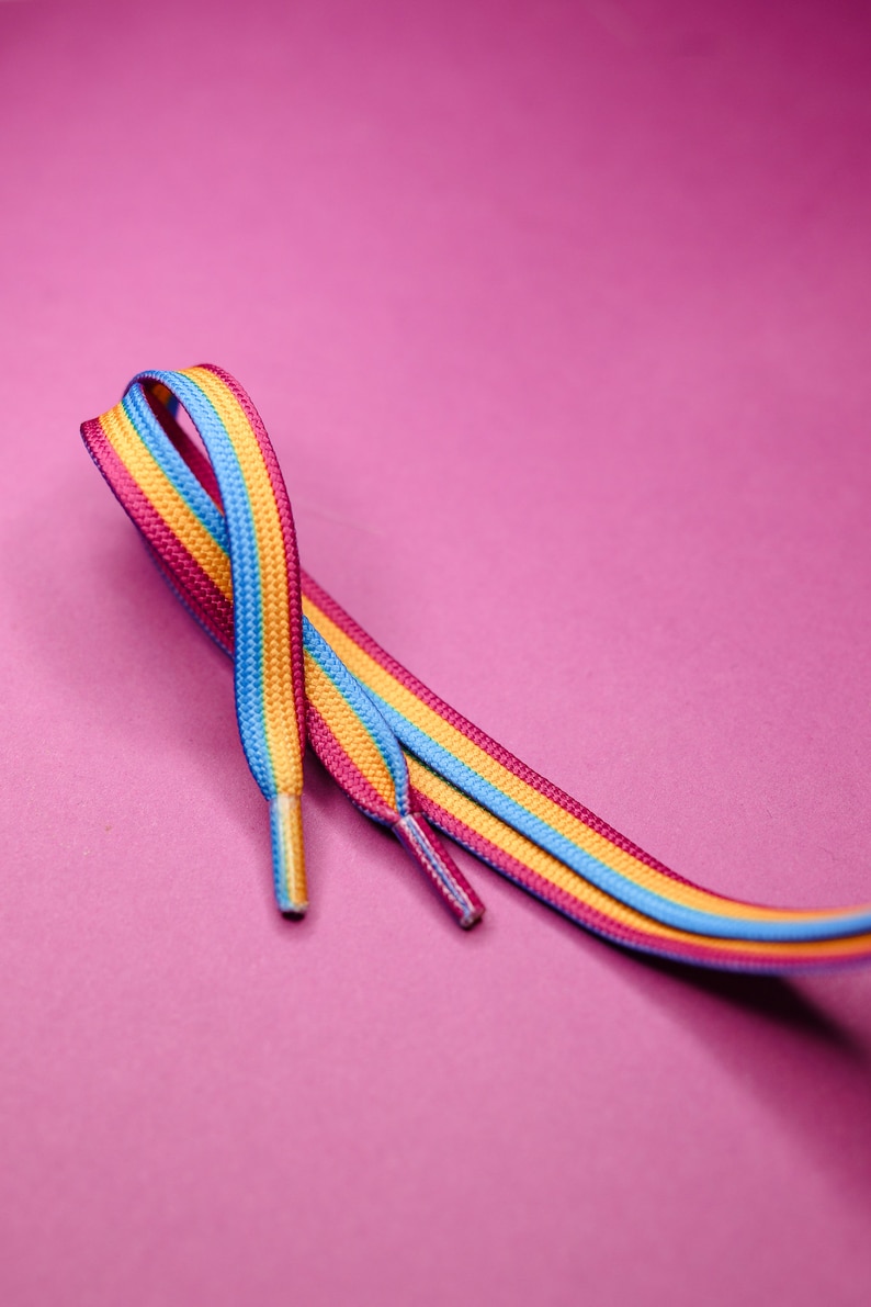 Pode incluir: Um par de atacadores listrados em rosa, amarelo e azul. Os atacadores s&atilde;o exibidos sobre um fundo rosa. Um acess&oacute;rio vibrante.