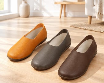 May include: Three slip-on shoes with rounded toes and textured surfaces, arranged in a row. The shoes are coloured in shades of mustard yellow, slate grey, and dark brown, each featuring a light-coloured interior lining and a flat sole.