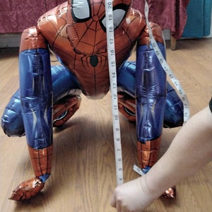 May include: A red, blue, and black Spider-Man balloon, approximately 24 inches tall, is shown on a wooden floor. The balloon is in a crawling position, with a tape measure held up to it to measure its height. The balloon has the Spider-Man logo on the chest.
