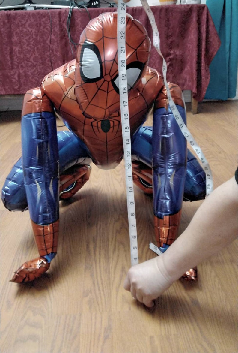 May include: A red, blue, and black Spider-Man balloon, approximately 24 inches tall, is shown on a wooden floor. The balloon is in a crawling position, with a tape measure held up to it to measure its height. The balloon has the Spider-Man logo on the chest.