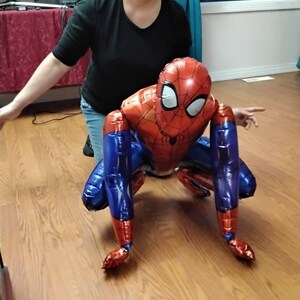 May include: A red, blue, and white Spider-Man balloon in a crouching position. The balloon has a detailed design of the superhero's suit, including the mask and web pattern. The balloon is on a wooden floor.